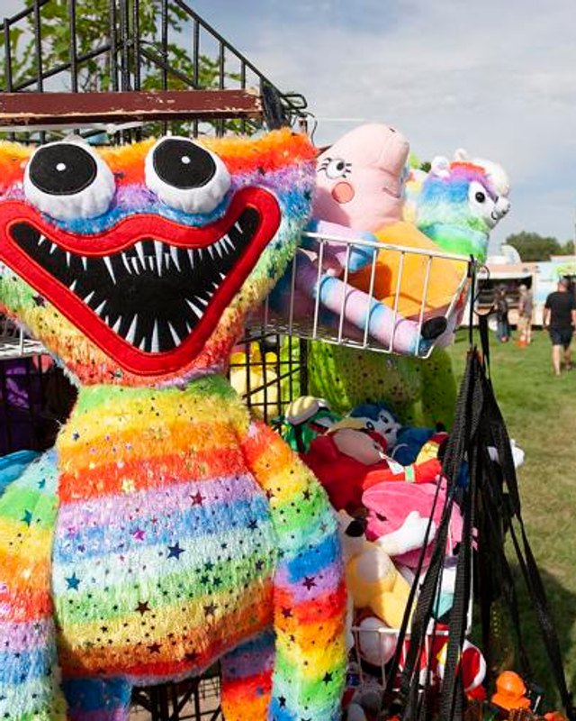 Colorful plush toy display and a crowd at a Kuna Days festival booth on the grass in Bernie Fisher Park.