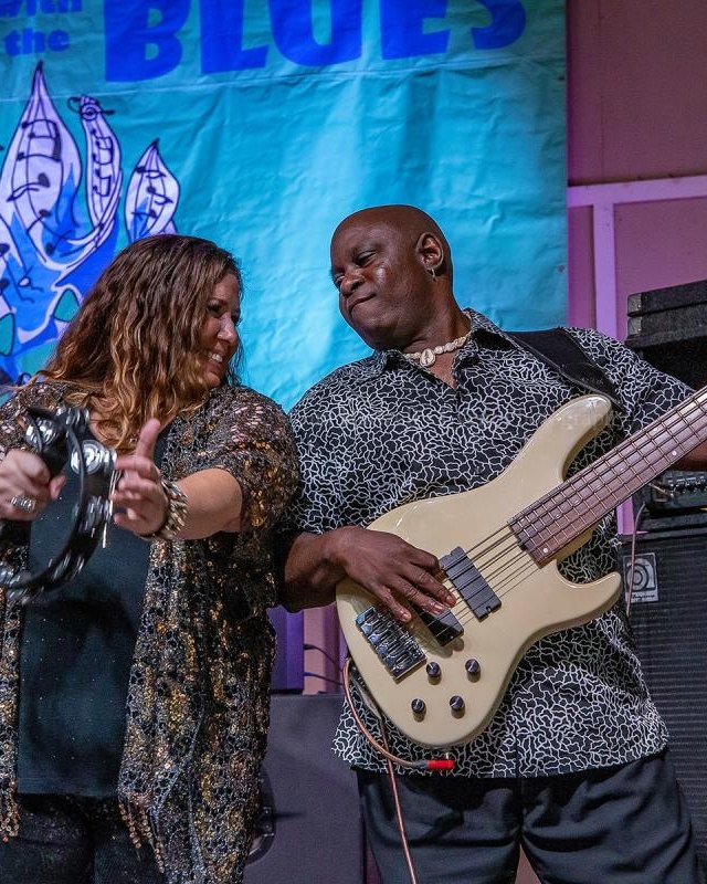 Musicians performing at Camping with the Blues: a woman holding a tambourine beside a bassist playing a bass guitar, in front of a blue banner.