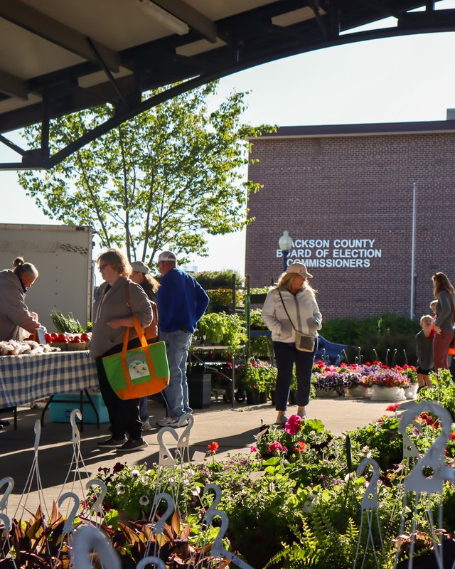 Shoppers and vendors at market tables with flowers under a covered pavilion at Independence Uptown Market.