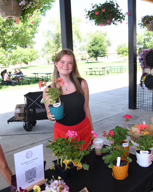 A smiling young vendor holds a potted geranium at a flower booth with pink blooms under a park pavilion in Windsor.