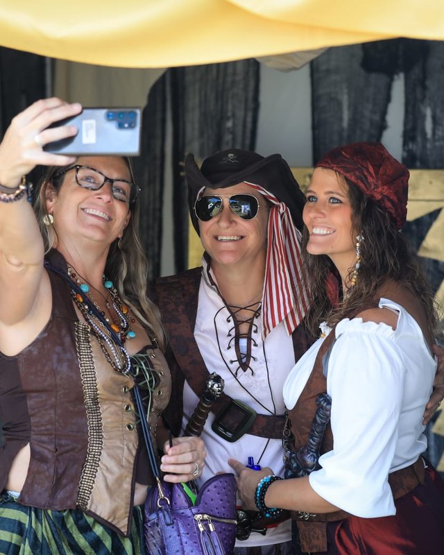 Three costumed pirate women posing and taking a selfie at a pirate-themed tent at Boynton Beach Pirate Fest.