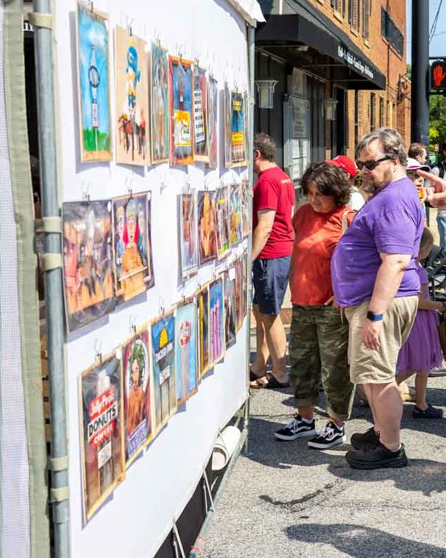Two visitors in red and purple shirts looking at art prints hung on a street-side display panel at an outdoor art festival.