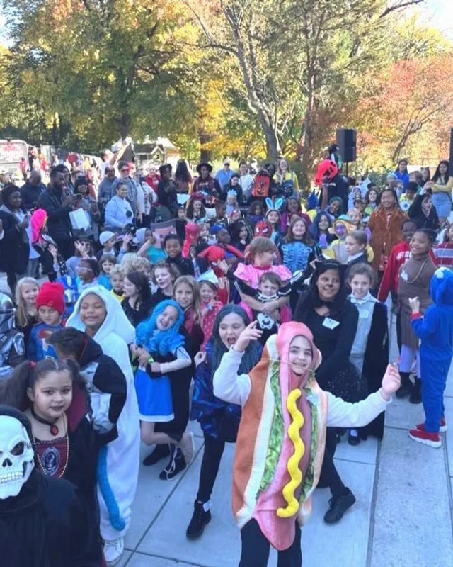 Crowd of children and adults in Halloween costumes gathered outdoors at a community event.