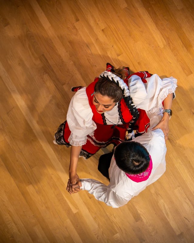 Two folkloric dancers seen from above on a wooden floor: one in a white and red dress, one in a white shirt with a pink bandana.