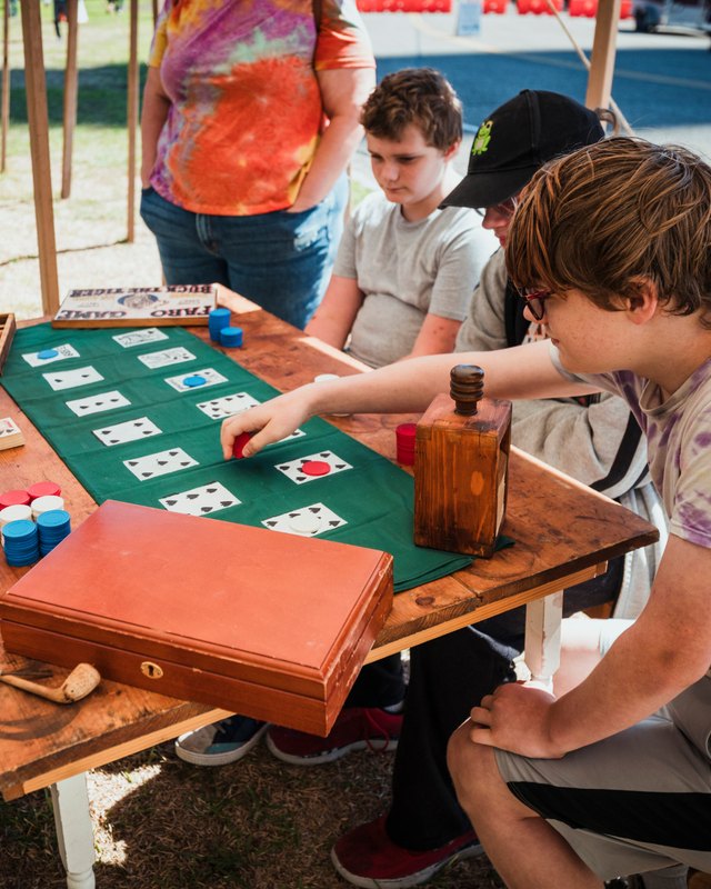 Teens play a card game on a wooden table with green felt at the San Jacinto Fall Fandango, with an adult standing behind them.