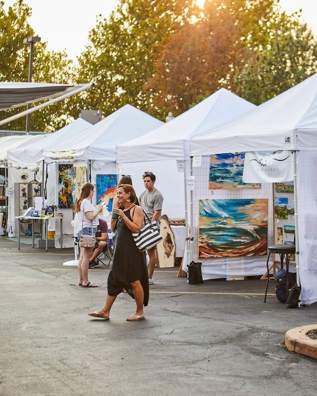 White tent booths with paintings on display and visitors walking through an outdoor art market