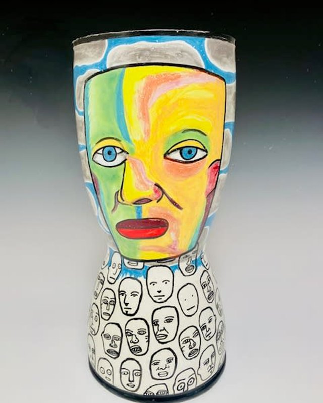 Painted clay vessel with a yellow, green and blue face and rows of black-and-white faces at the base, on a dark gradient background.