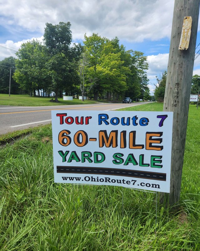 Roadside sign reading Tour Route 7 60-Mile Yard Sale on a utility pole beside a grassy road.