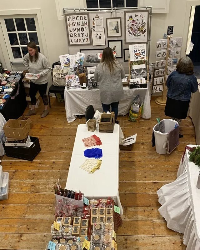 Shoppers browsing craft vendor booths with handmade goods and wreaths inside a wood-floored hall