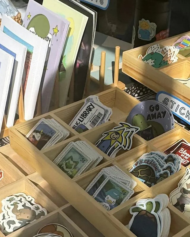 Stickers and prints displayed in wooden compartment trays at a craft market booth.