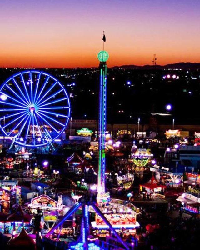 Night view of the Arizona State Fair midway with a lit blue Ferris wheel and tall tower ride over the crowds.