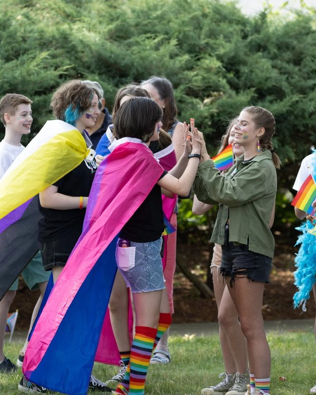Festival-goers in rainbow capes and flags gathering and greeting each other on a green lawn.