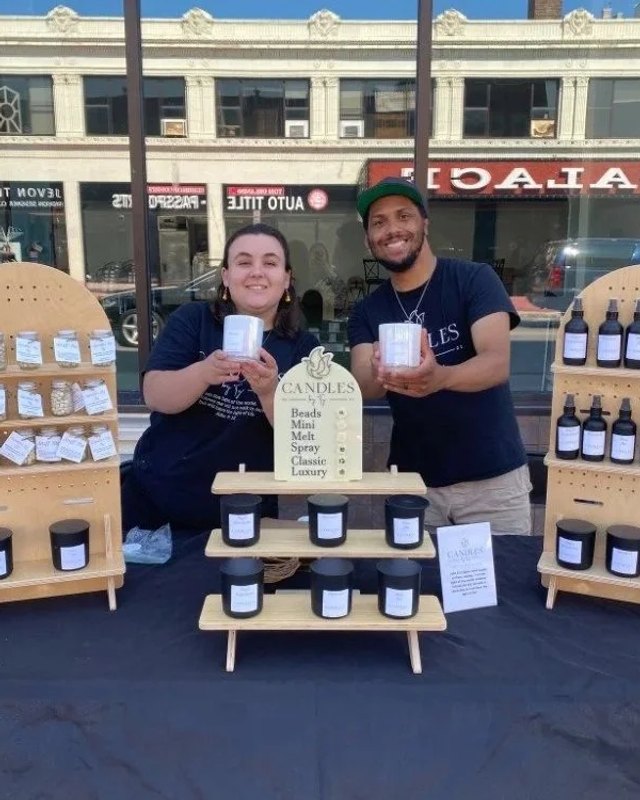 Two vendors behind a display of candles and bottled products at a Second Saturdays booth.