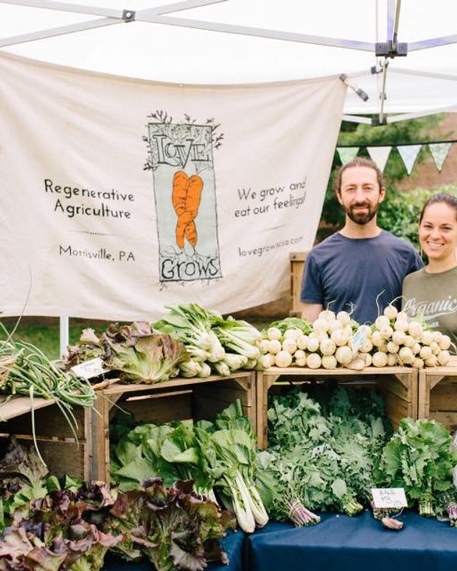 Produce stall with crates of greens and root vegetables under a white tent, with two farmers behind a Regenerative Agriculture banner.