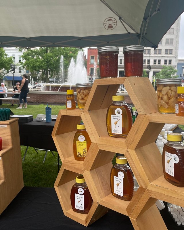 Honey jars and candles displayed on hexagonal wooden shelves at an outdoor market booth with a fountain behind.