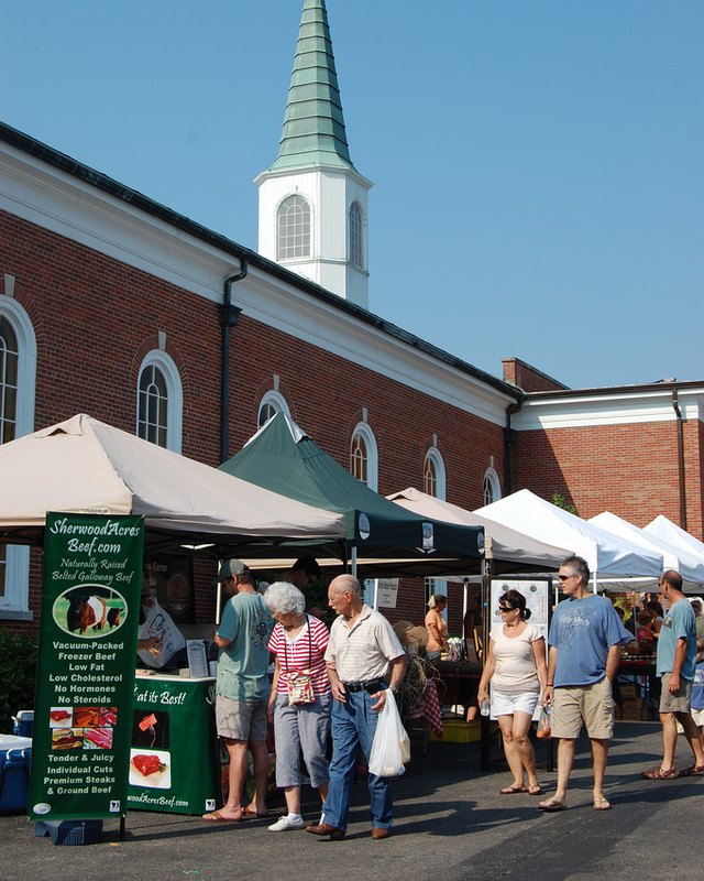 Shoppers and vendor tents at the St. Matthews Farmers Market beside a brick church with a steeple.