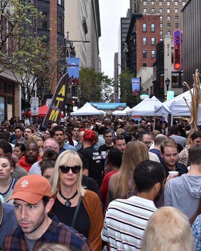 Dense crowd walking along a city street past white vendor tents at the Midtown Village festival