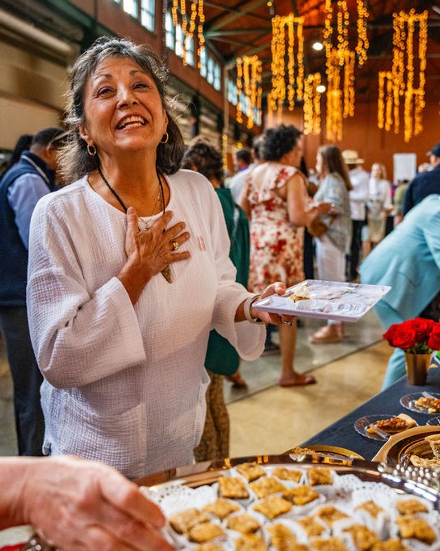 A smiling woman holds a tray of appetizers at an indoor event with string lights and a crowd behind her.