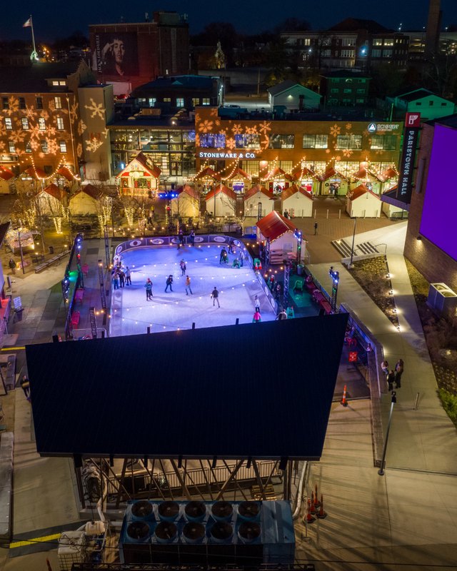 Aerial night view of the Fête de Noël holiday market in Paristown, with lit stalls, an outdoor skating rink and a large dark screen.
