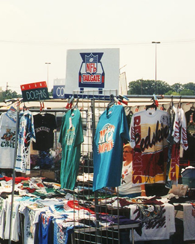 Clothing and T-shirt vendor booths with racks of shirts under a sign at the Philadelphia Flea Market.