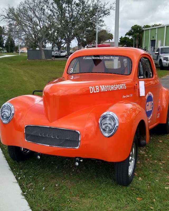 Bright orange vintage hot rod coupe with DLB Motorsports lettering parked on grass at a car show.
