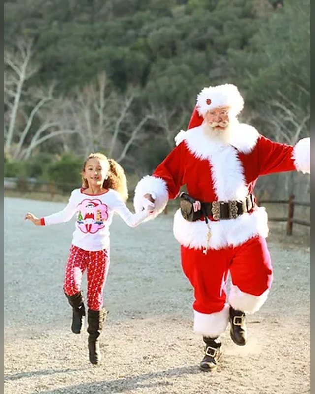 Santa in a red suit running with a young girl on a dirt path outdoors.